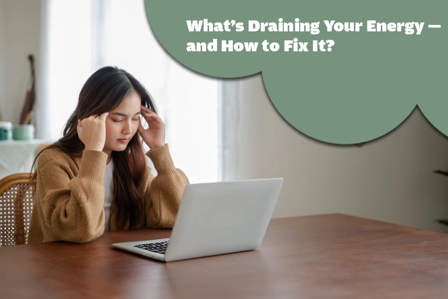 A woman sitting at a table using a laptop while holding her head, connected to energy drain and mental fatigue.